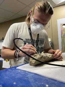 Spencer Shroyer wears protective glasses and mask as he works to extract fossil from rock