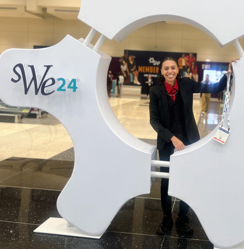 Young woman smiles in front of gear display with words SWE24 (Society of Women Engineers) in lobby