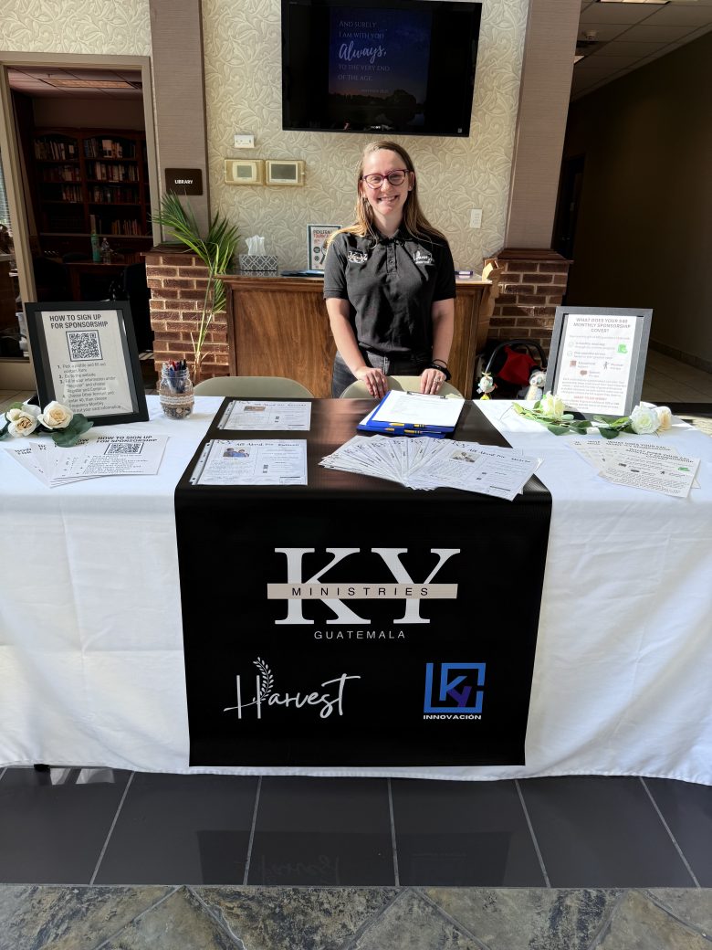 A smiling woman stands behind a table promoting KY Ministries Guatemala, with brochures, sign-up sheets, and informational displays set up