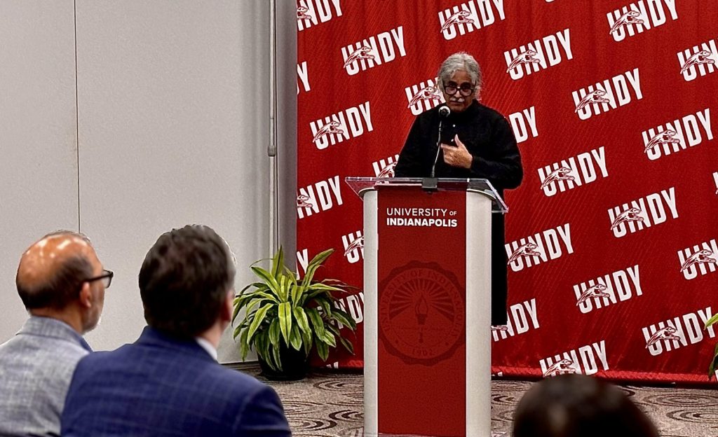 Man stands at podium with microphone in lecture hall in front of red UIndy backdrop. Three audience members in foreground 