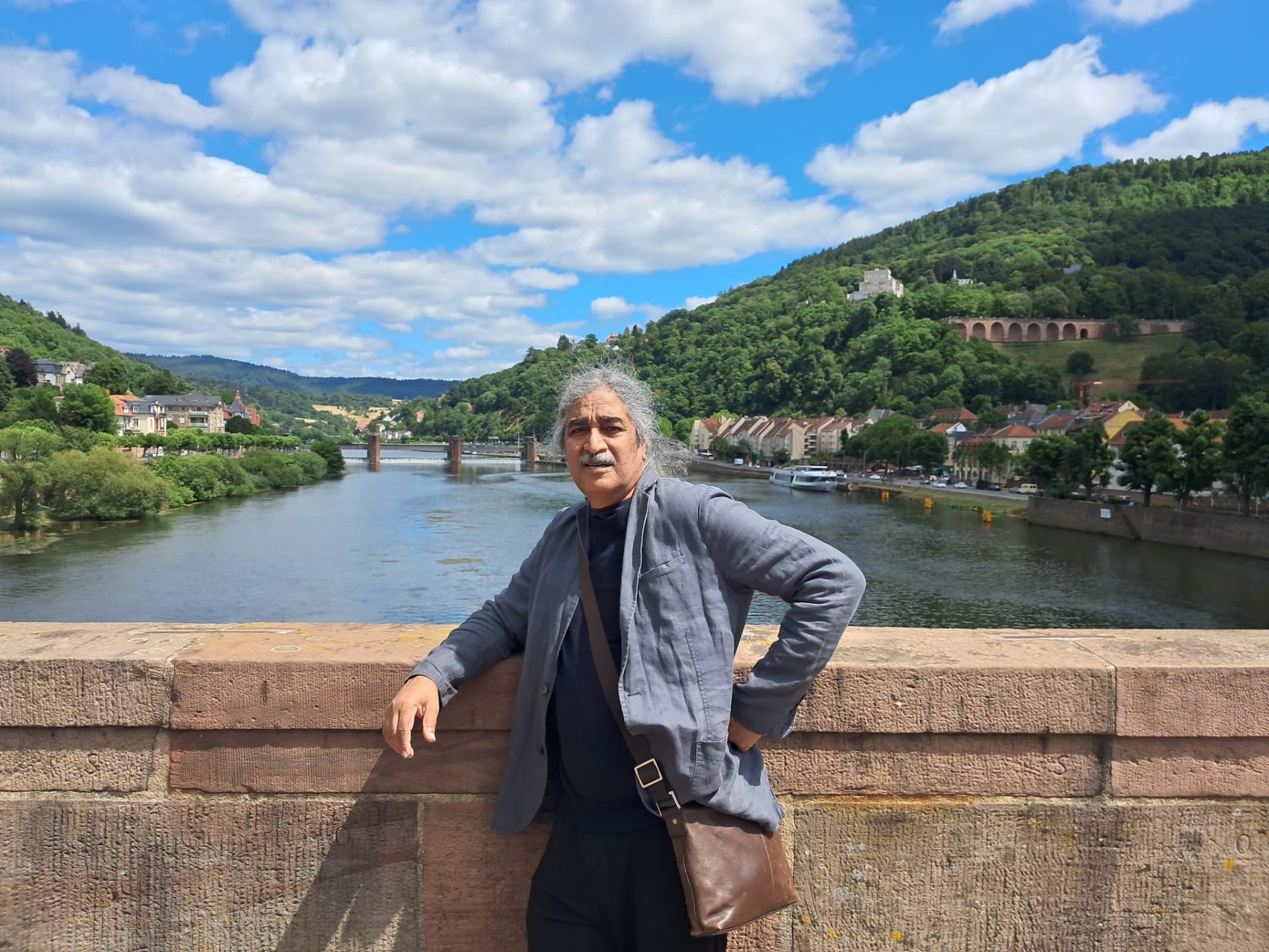 Man stands at riverfront, with scenic landscape with green picturesque trees and blue skies