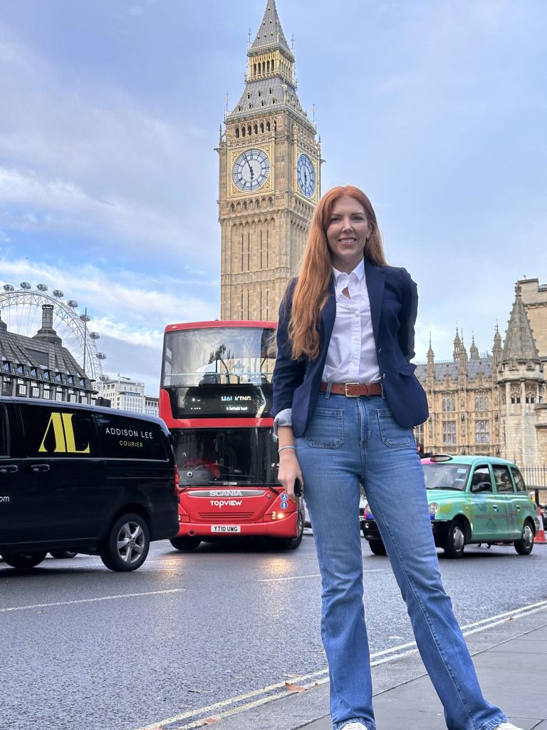 Woman in business casual attire, stands in front of Big Ben in England with cars, busses in background
