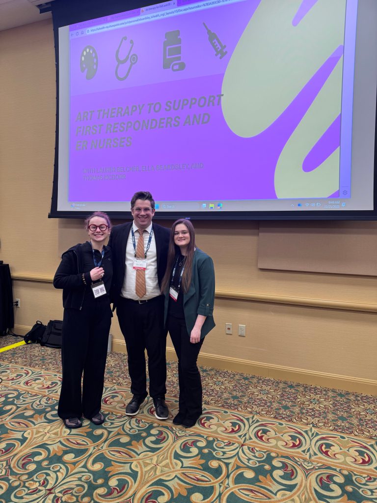 Three presenters stand smiling in front of a projected presentation titled "Art Therapy to Support First Responders and ER Nurses" at a conference. The screen behind them displays art, healthcare, medication, and syringe icons, and each presenter is wearing a conference name badge.