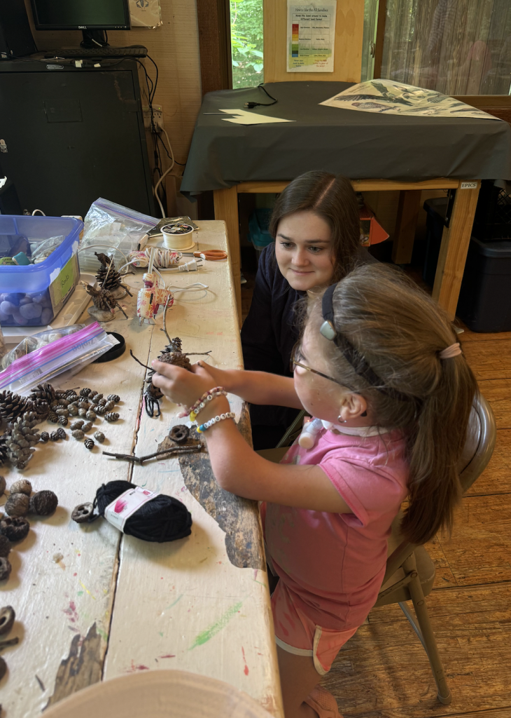 A facilitator sits beside a young child at a craft table covered with pinecones, sticks, yarn, and other natural materials.