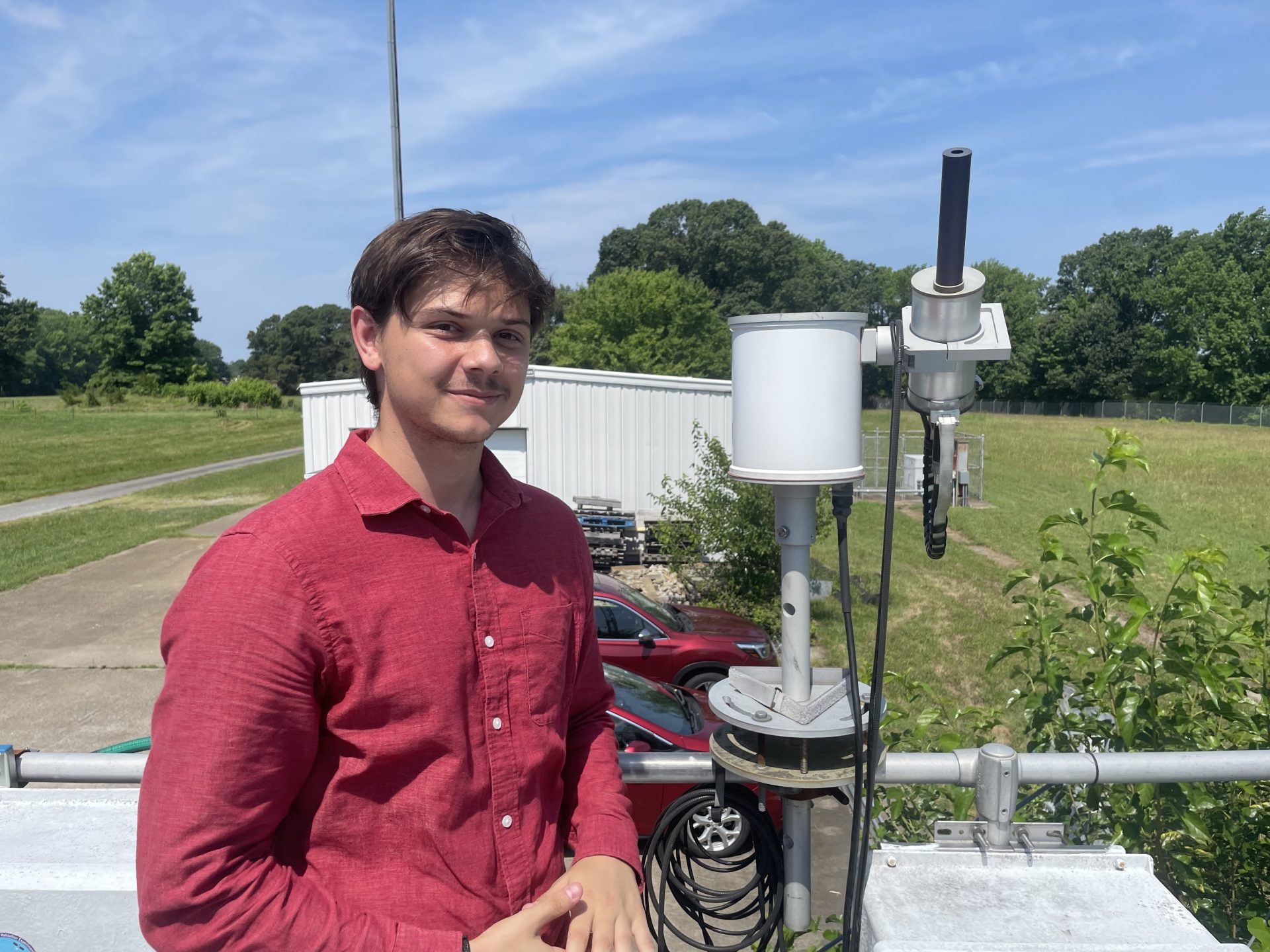 Man in red dress shirt smiles next to viewing device with field in background