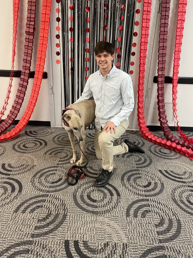 Man in dress clothes kneels next to greyhound mascot in ballroom with red decorations in background 